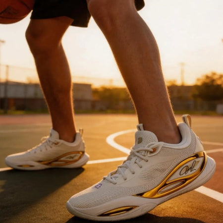 A person standing on an outdoor basketball court at sunset wearing white athletic sneakers with gold and black accents. The photo is cropped from the knees down, with a basketball partially visible on the left.