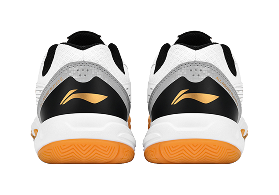 Pair of white badminton shoes with orange outsoles, black heel panels and gold logo