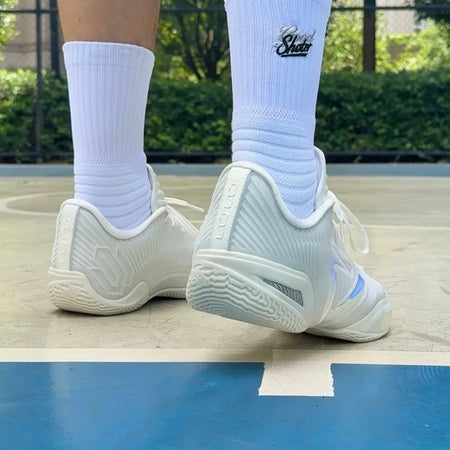 White athletic socks and white sneakers worn by a person standing on an outdoor basketball court, with a blue court surface visible below.
