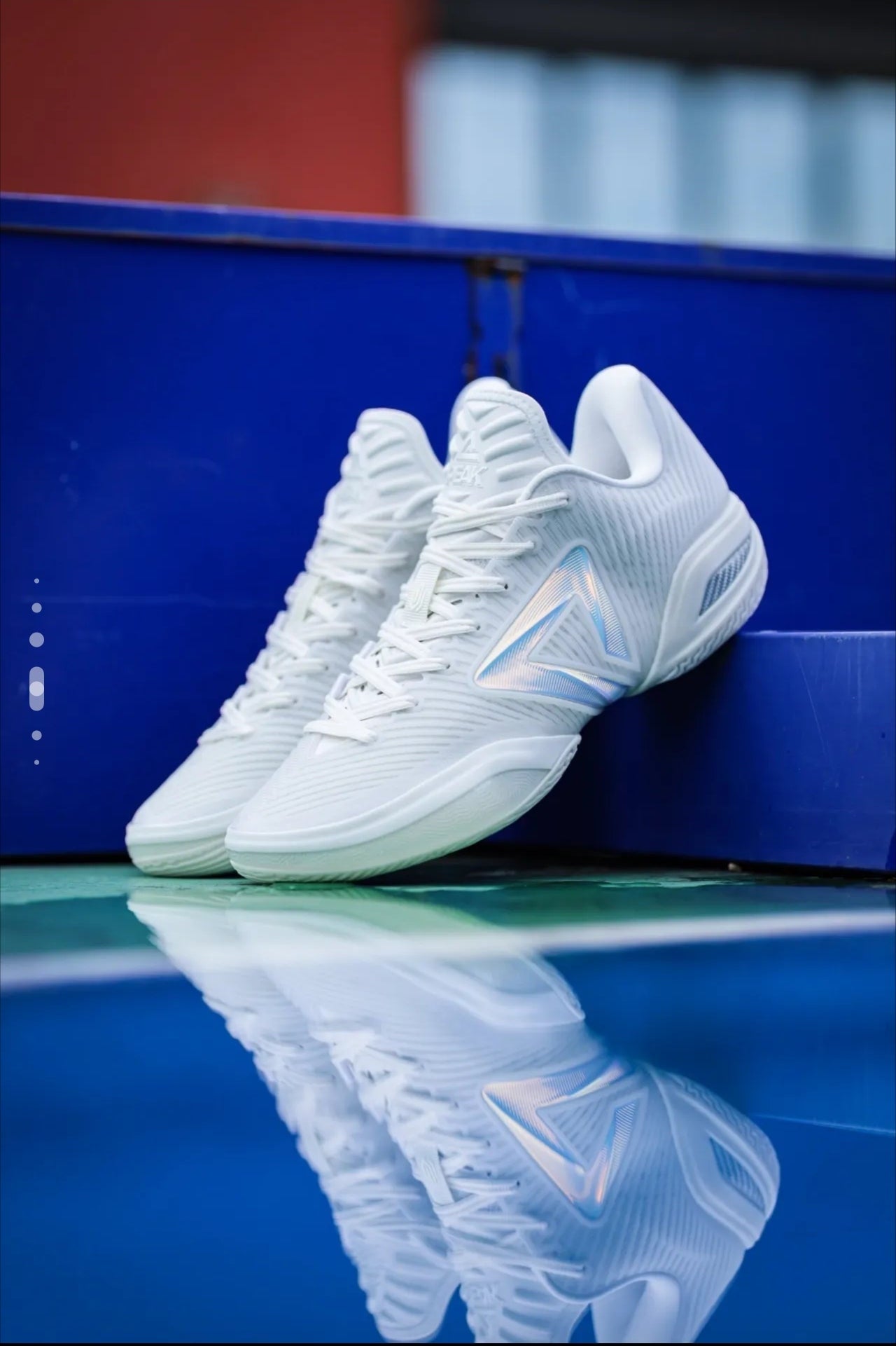 A pair of white athletic sneakers with subtle light blue accents, displayed on a reflective dark blue surface with a blue background.