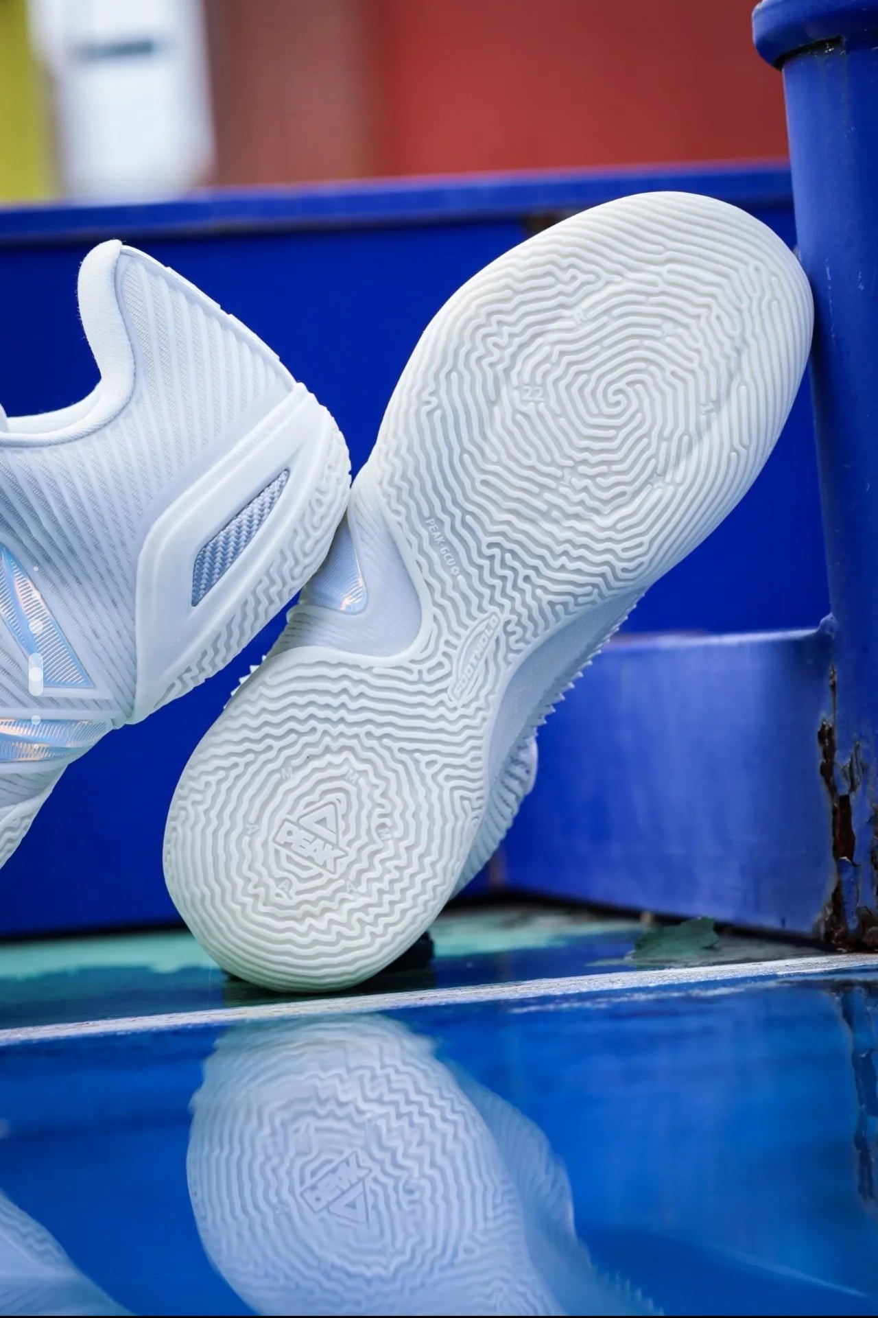 White athletic sneakers shown from the sole side, photographed outdoors against a blue structure with a reflective surface below.
