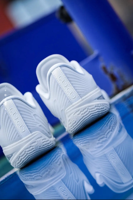 A pair of white low-top sneakers with textured wavy patterns, displayed on a reflective blue surface.