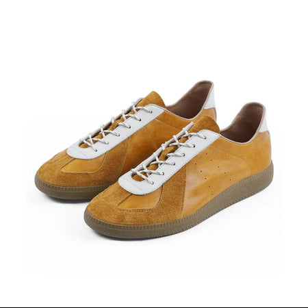 A pair of low-top suede sneakers in mustard yellow and brown, featuring white laces with gold tips and white side stripes, shown on a white background within a mobile app screenshot.