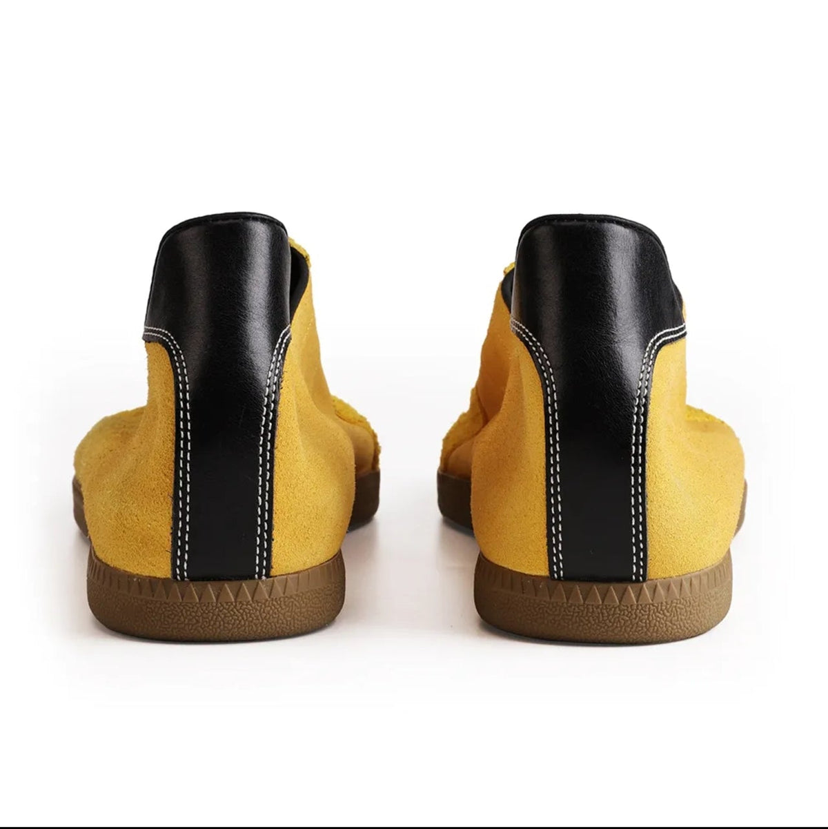 A pair of yellow suede ankle boots with black leather heel panels and brown gum soles, shown from the back against a white background.