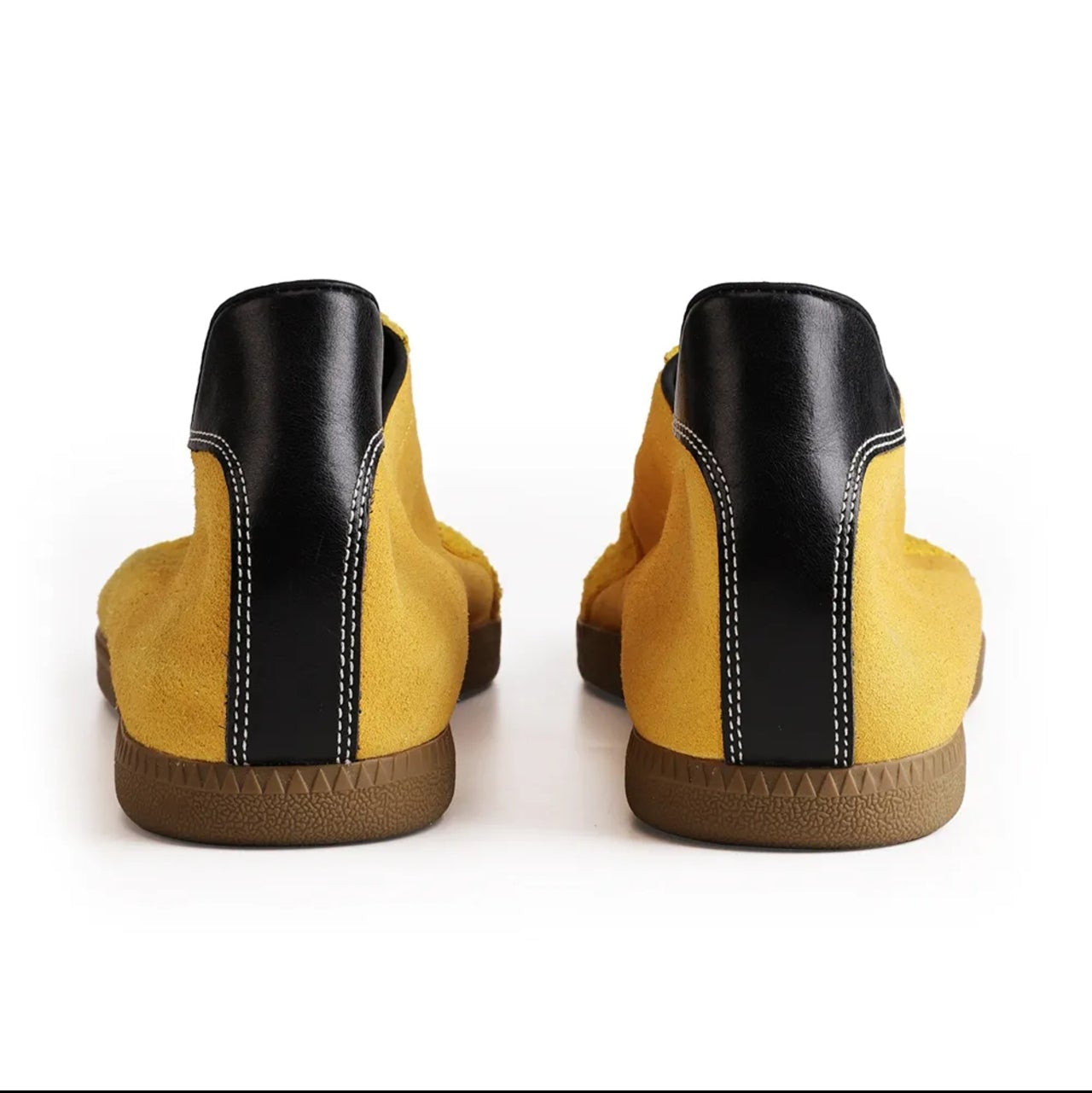 A pair of yellow suede ankle boots with black leather heel panels and brown gum soles, shown from the back against a white background.